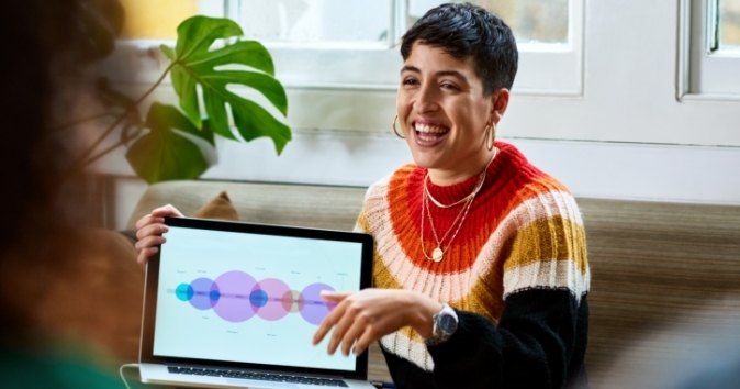 A woman with short hair is smiling and gesturing while presenting in a casual setting. She is holding a laptop that displays a colorful diagram with overlapping circles and text. The background features a large green plant and windows, creating a bright atmosphere.