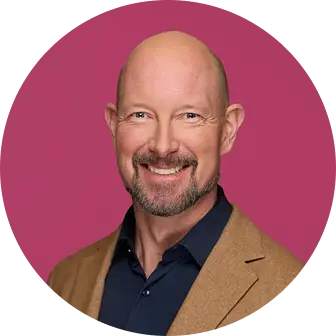 A smiling man with a bald head and a well-groomed beard is dressed in a navy shirt and a brown blazer, set against a bright pink background.