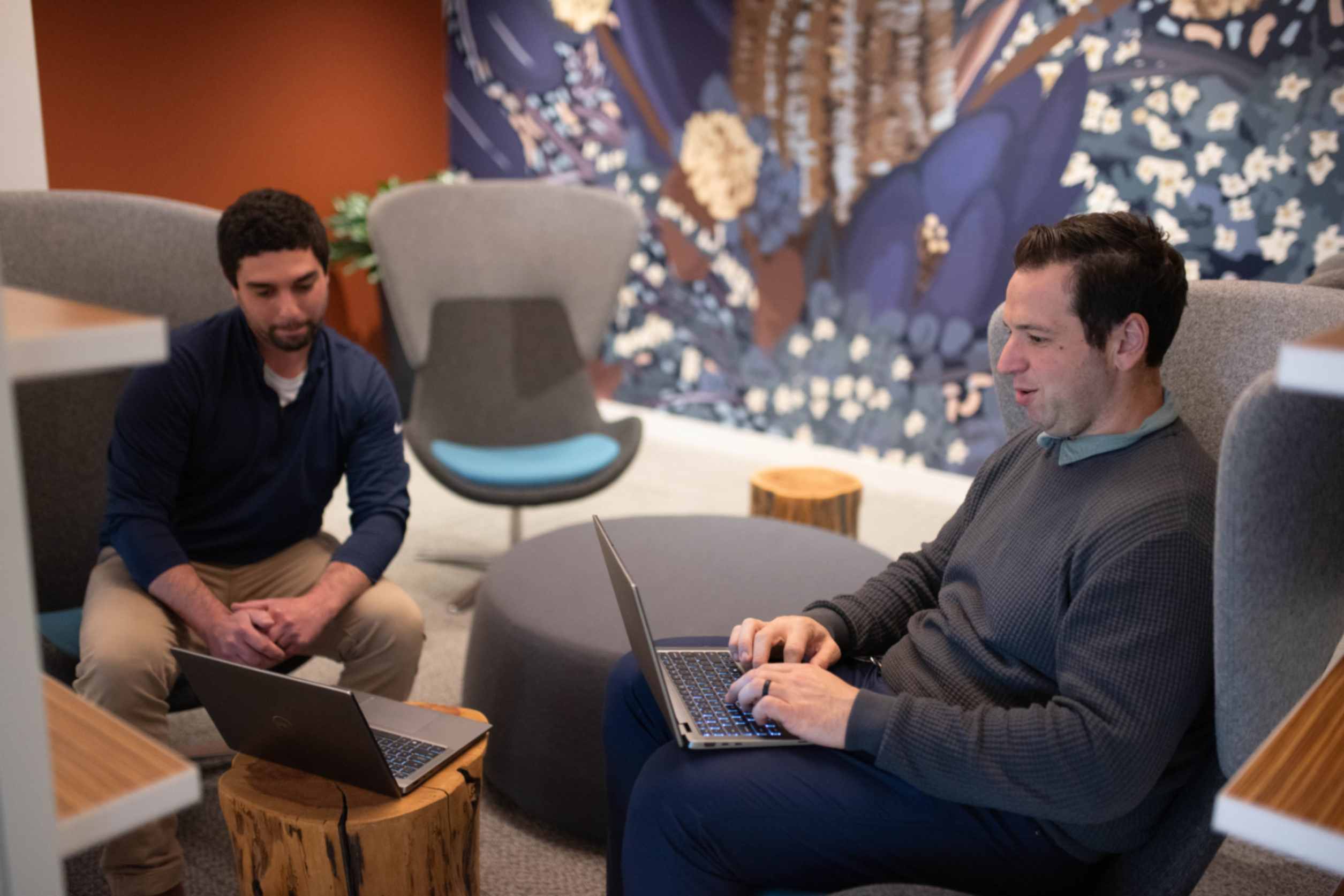 Two men are seated in a modern workspace with a colorful mural in the background. One man, wearing a navy blue shirt, is sitting on a stool with his hands clasped, while the other man, in a grey sweater, is working on a laptop placed on his lap. They are engaged in conversation in a comfortable setting that includes a round, grey table and a decorative chair.