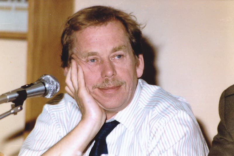 Václav Havel, with a mustache rests his chin on his hand, sitting beside a microphone during a discussion. He wears a striped shirt and a dark tie, displaying a thoughtful expression. The background features a simple interior setting.