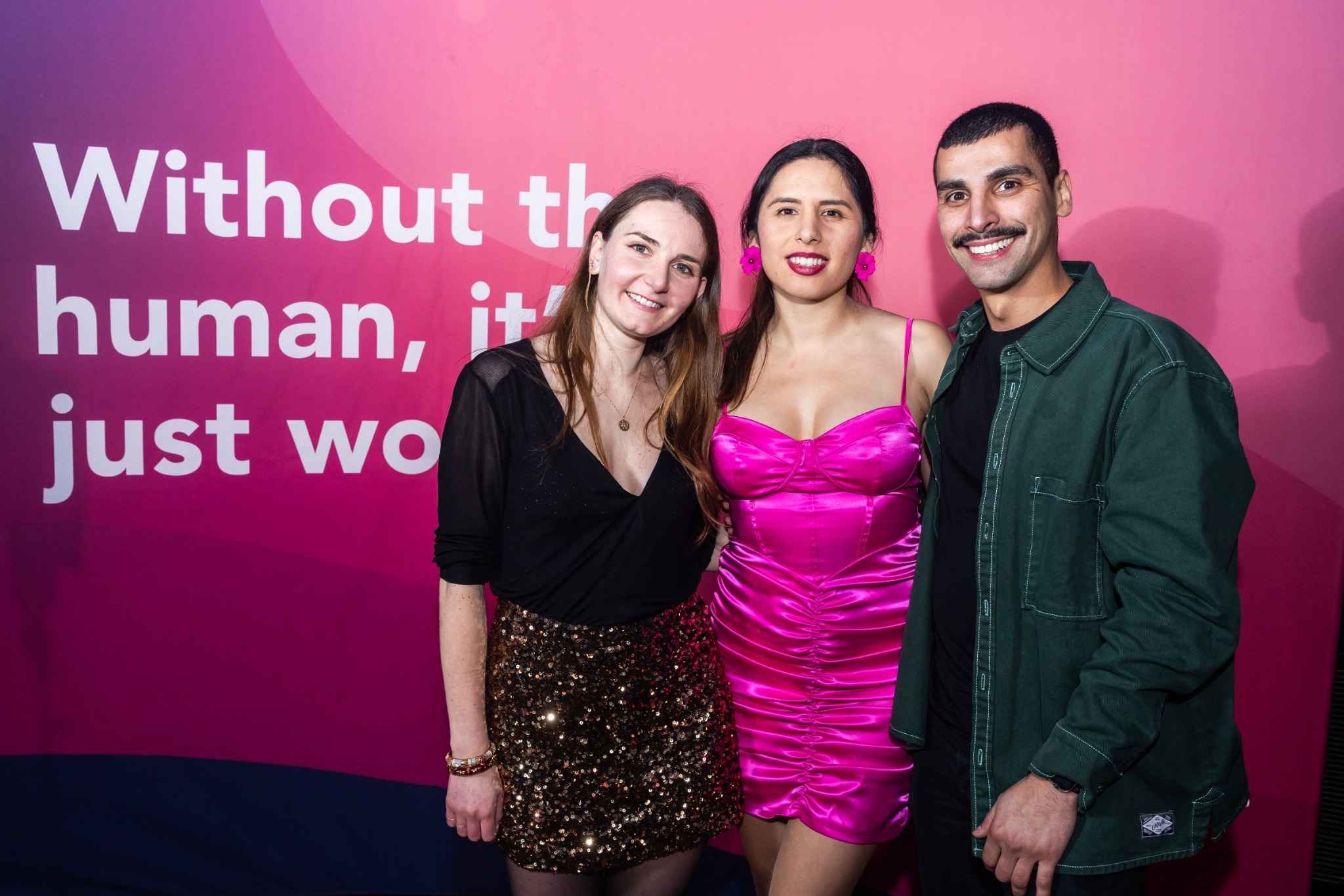 Three people pose smiling in front of a vibrant pink backdrop that reads "Without the human, it's just work." The person on the left wears a black top and sequined skirt, the person in the center wears a bright pink dress, and the person on the right wears a green jacket over a black shirt