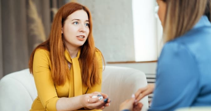 A woman with long red hair is sitting on a white couch, engaging in conversation with another woman whose back is facing the viewer. The woman in yellow is animatedly speaking while holding a small object in her hands. The setting appears modern and well-lit, with soft furnishings and a neutral background.