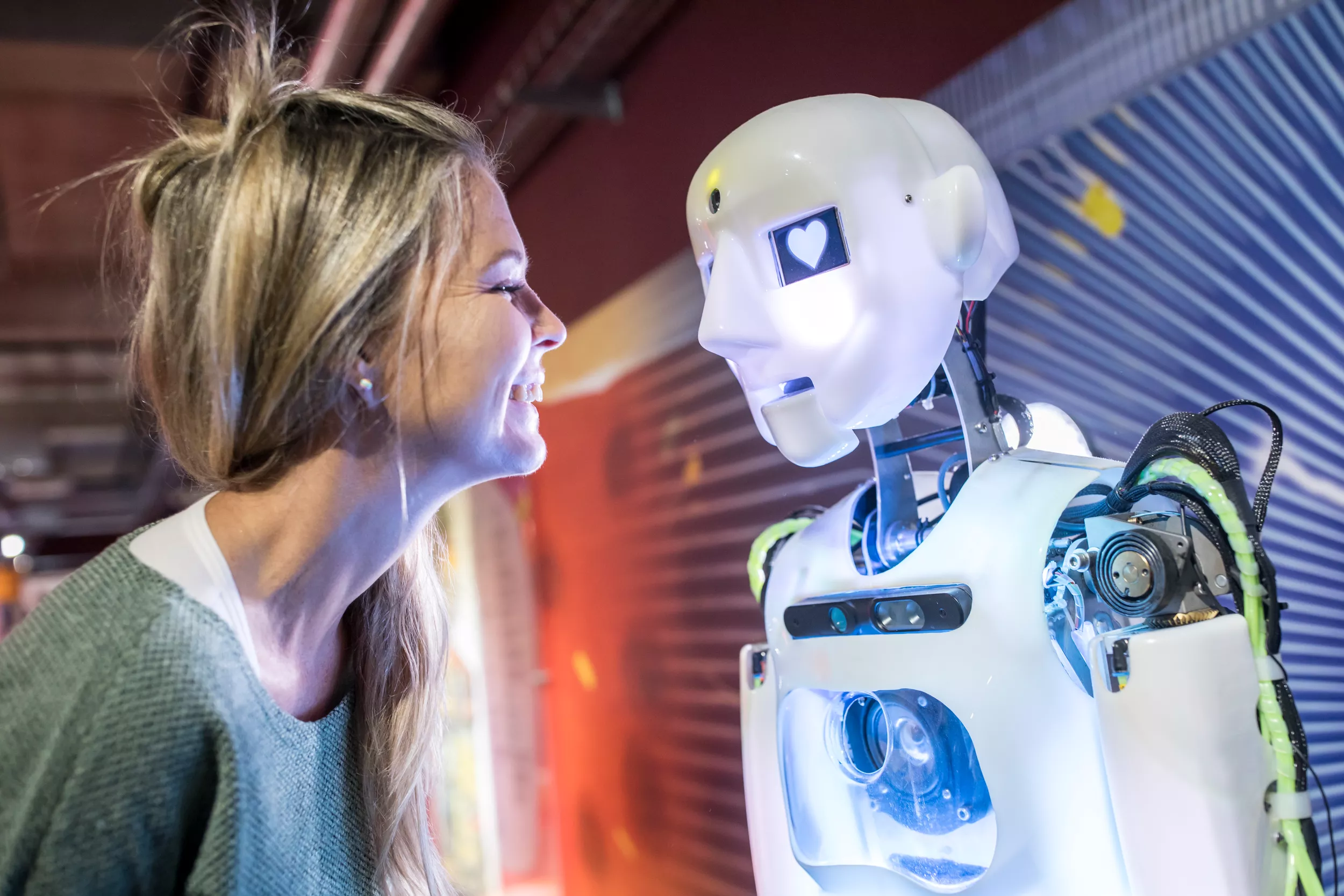 A woman with long, straight hair is smiling broadly while leaning towards a robot. The robot has a simple face with an illuminated heart-shaped display in place of its eyes. The background features colorful, abstract patterns, creating a lively setting for the interaction.