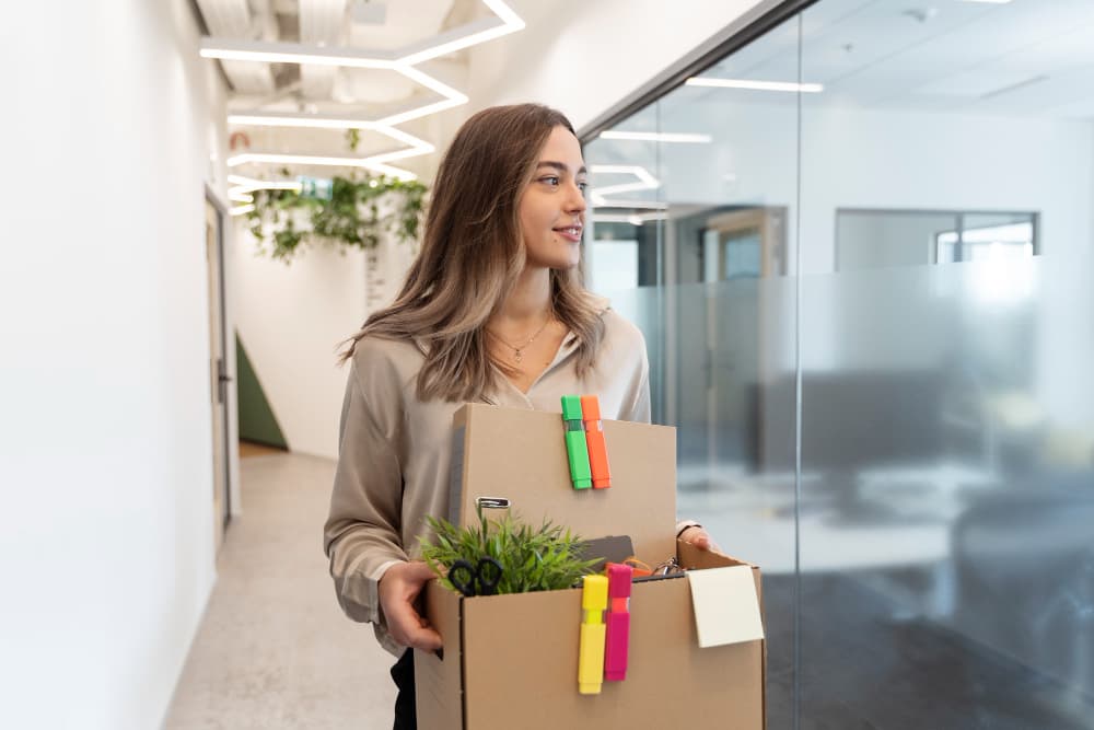 A woman is walking down a modern office corridor, carrying a box filled with office supplies, including scissors and a small potted plant. She has a pleasant smile on her face and is dressed in a light-colored blouse. The corridor features clean lines, greenery, and contemporary lighting, contributing to a bright and welcoming atmosphere.