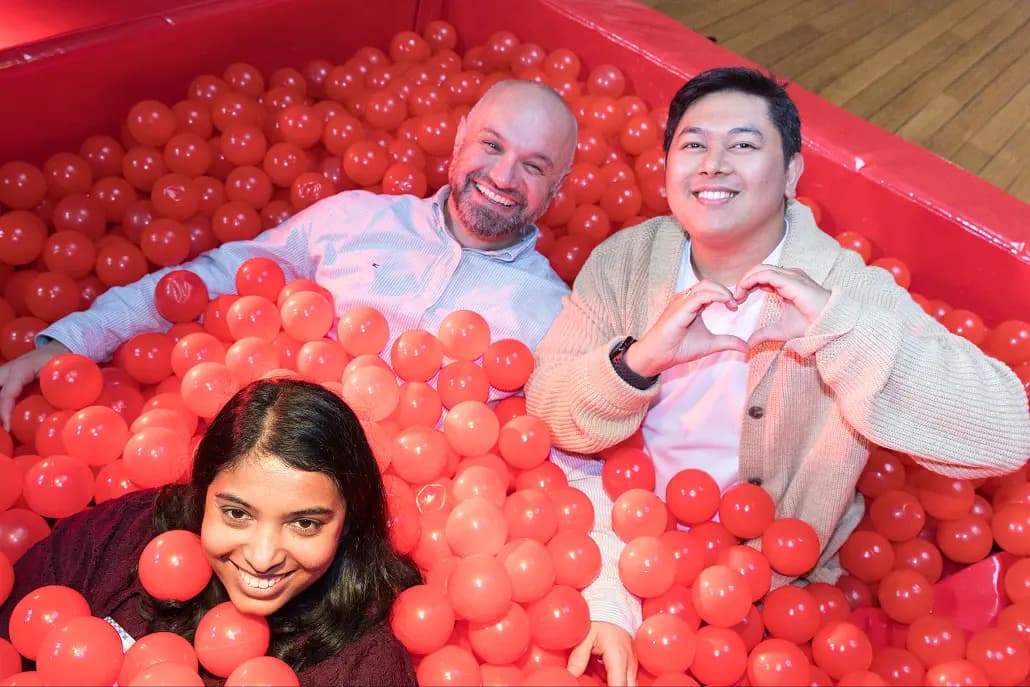 A woman and two men are playfully smiling while sitting among numerous red balls in a vibrant ball pit. One man is making a heart shape with his hands, evoking a sense of fun and camaraderie in the colorful environment.