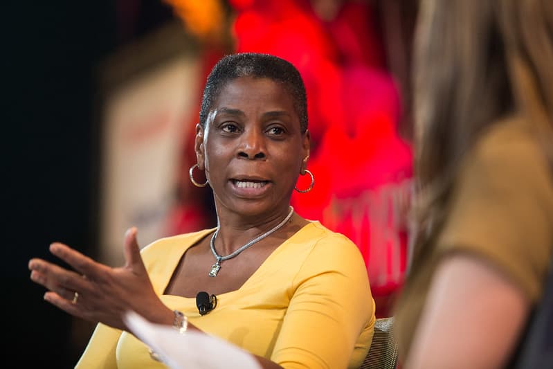 Ursula Burns, A woman in a yellow shirt is speaking passionately during a panel discussion. She has short hair and is gesturing with her hands to emphasize her points. In the background, colorful decorations and blurred text are visible, indicating a lively event atmosphere.