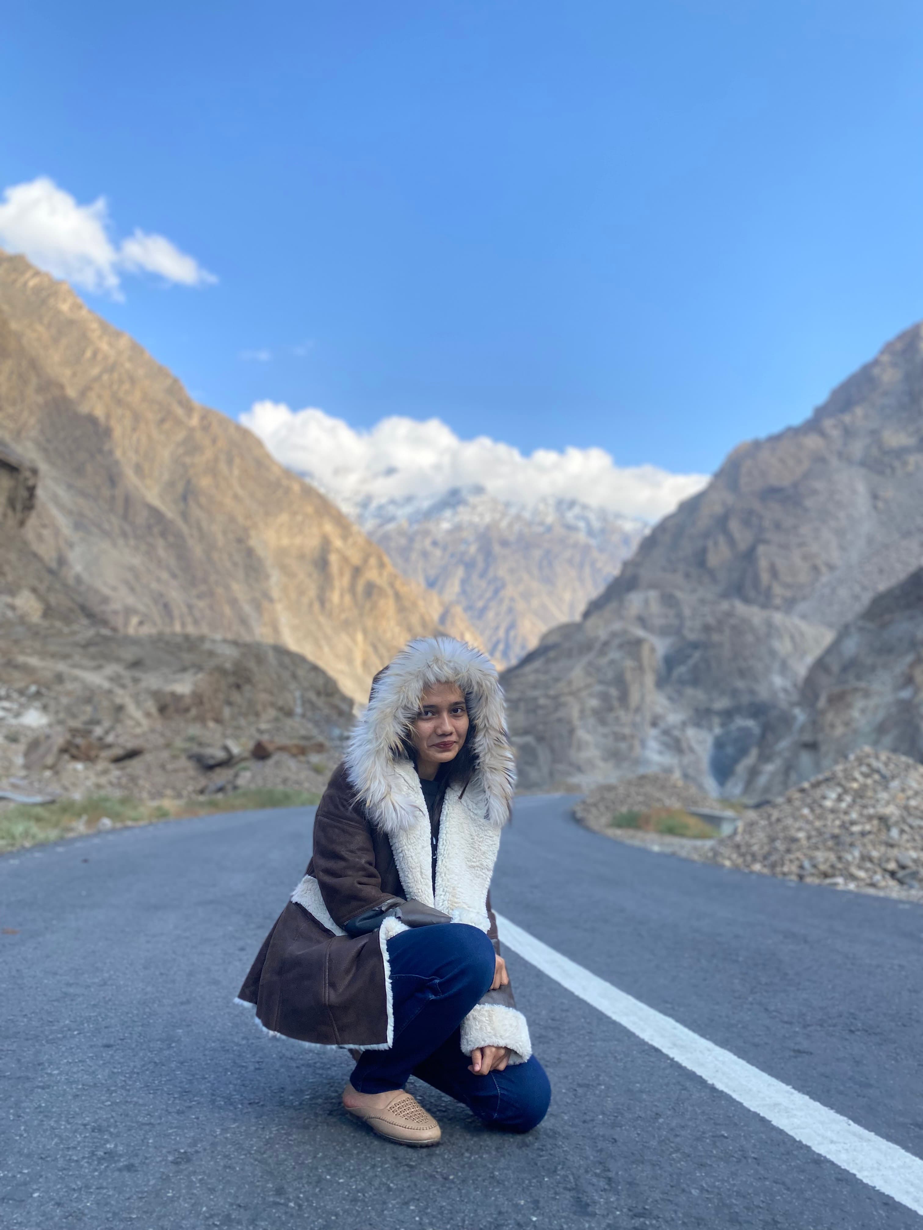 A person is kneeling on a winding road amidst a mountainous landscape. They are dressed in a warm coat with a fur-lined hood, and are surrounded by rocky terrain with mountains in the background under a clear blue sky with some clouds.