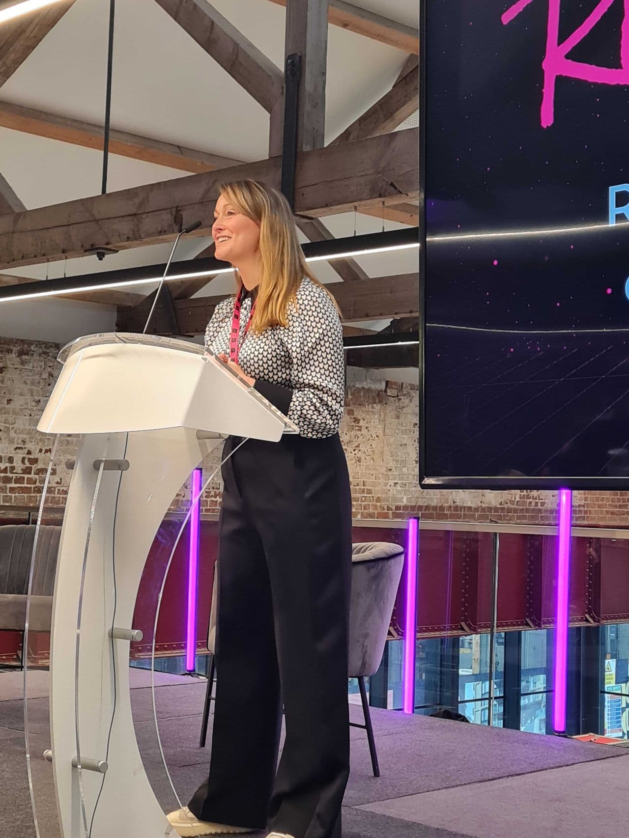 A woman with long hair is standing behind a podium, speaking at an event. She wears a patterned top and black pants, smiling as she addresses the audience. In the background, there is a large screen displaying the letters "K R," and the setting features wooden beams and a modern aesthetic. Two empty chairs are visible in the background, along with purple lighting accents.
