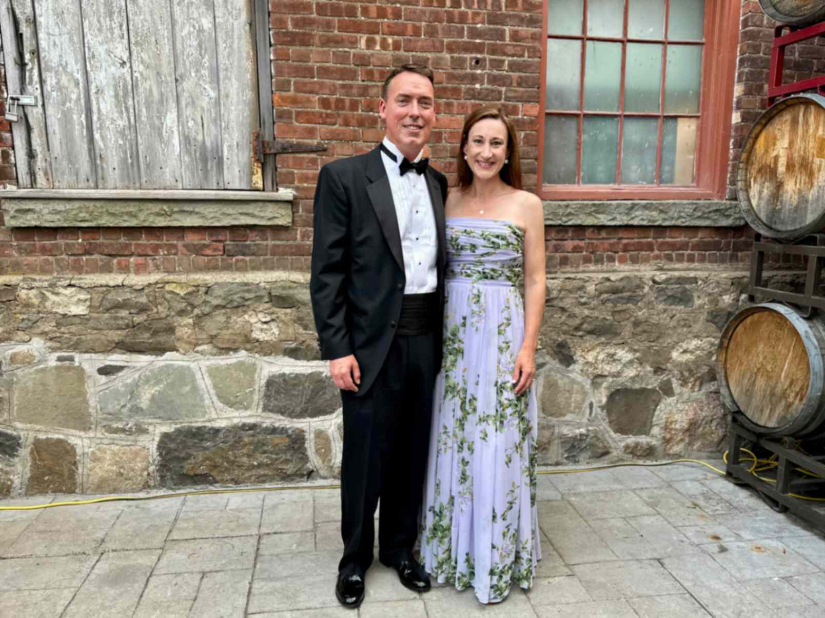 A couple stands in formal attire against a rustic brick wall with barrels. The man wears a black tuxedo, and the woman is in a purple floral gown. They are smiling, conveying a joyful tone.