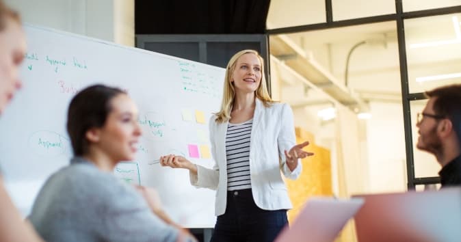 Woman talking at a white board to people in a room for a train the trainer session.