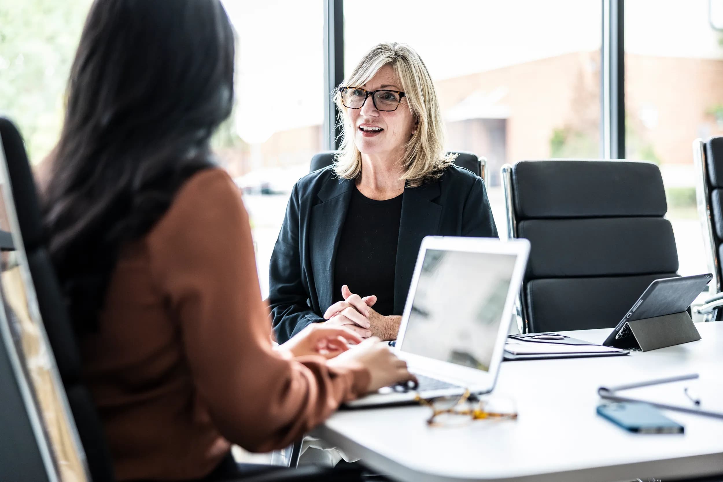 A woman with blonde hair and glasses is sitting at a conference table, engaged in conversation with another person whose back is visible. The setting has large windows with a view outside. On the table, there is a laptop and other office supplies. The woman appears animated and attentive, suggesting a collaborative discussion.