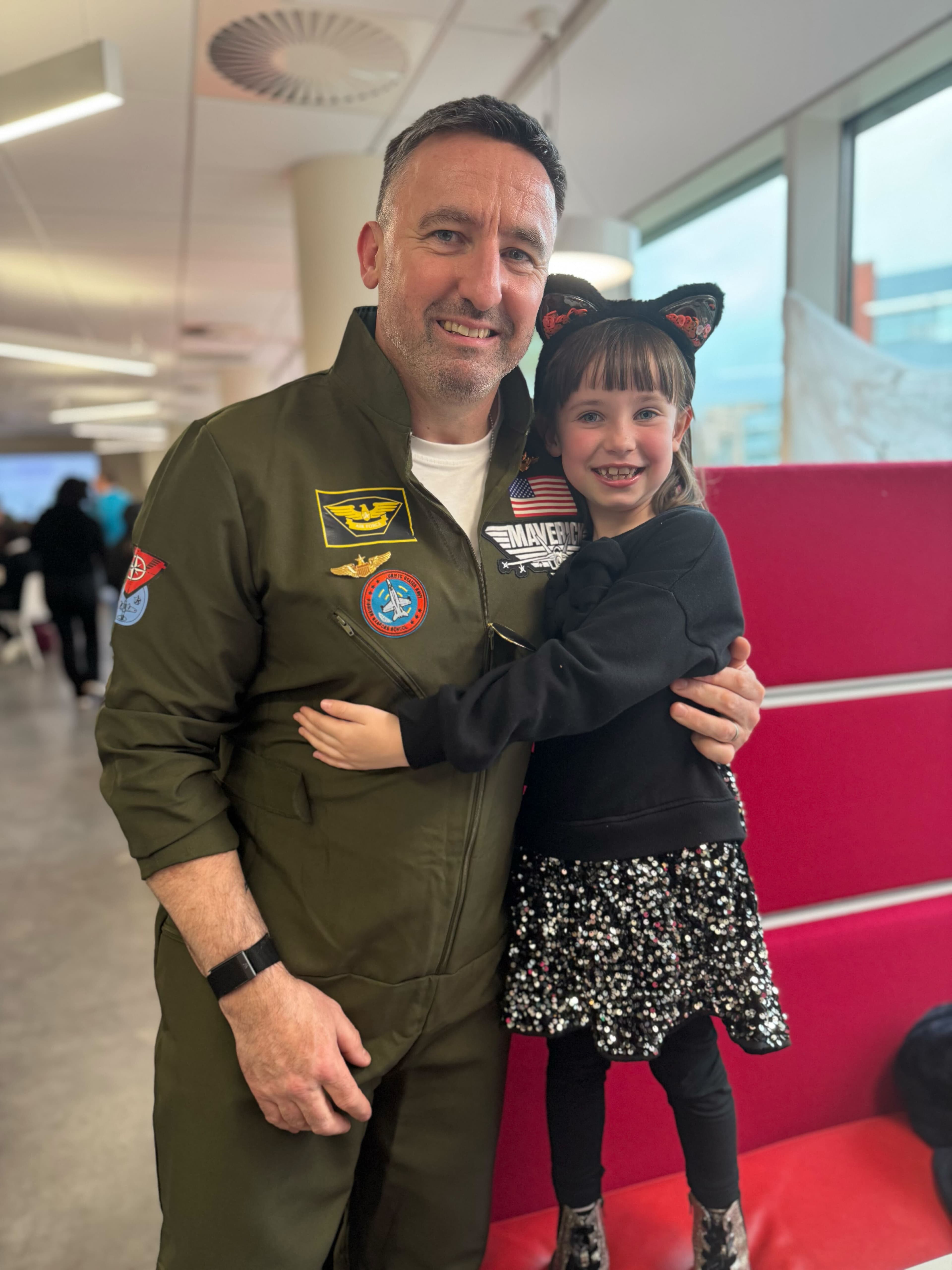 A man wearing a flight jacket smiles while holding a young girl in a black cat ear headband. The girl wears a black top and a sparkly skirt. They are in a bright, modern indoor space with large windows and colorful seating in the background.