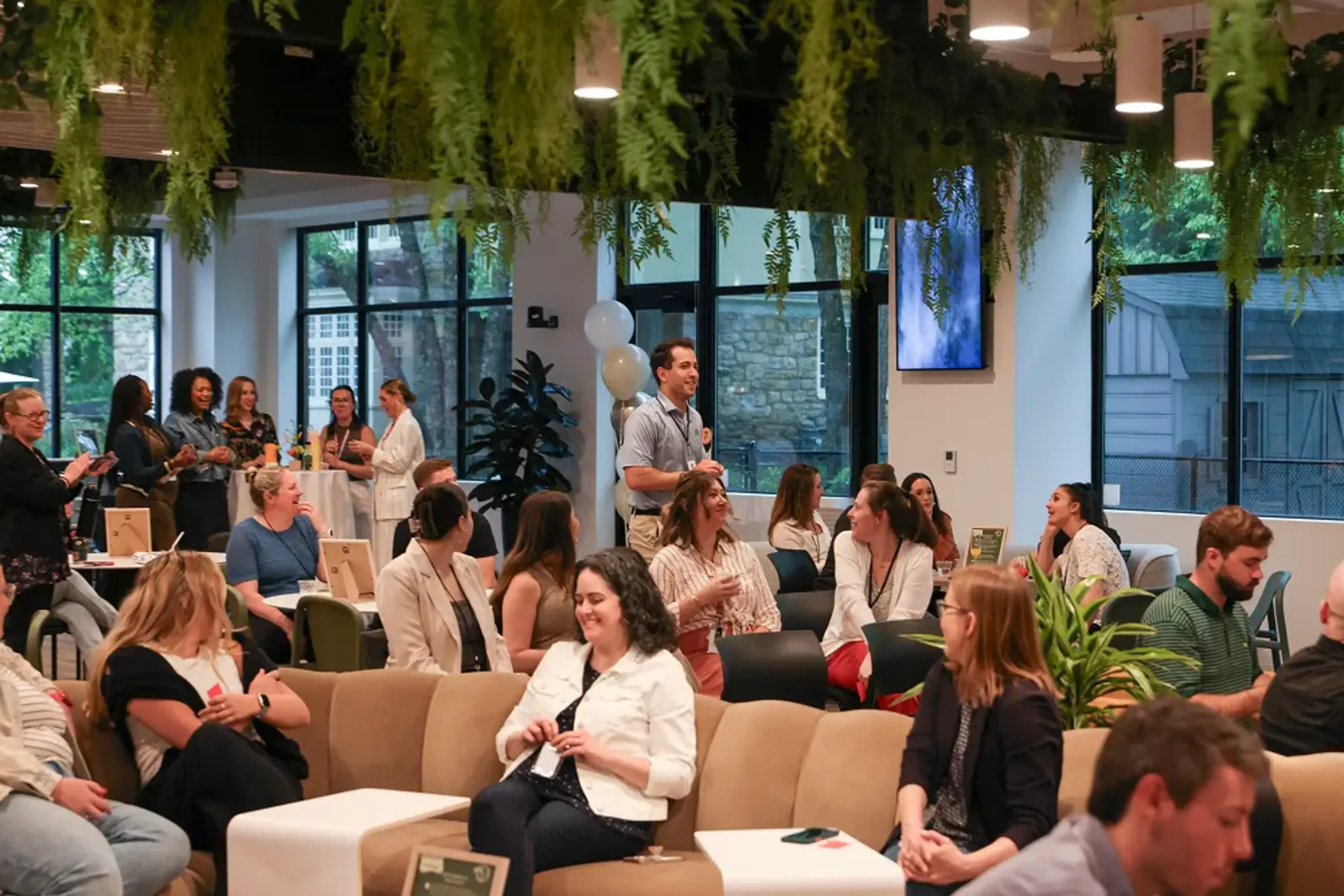 A lively indoor event scene featuring a diverse group of attendees engaged in conversation. In the foreground, several individuals are seated on a modern couch, some smiling and chatting. In the background, a few people stand near tables, while others mingle, with greenery hanging from the ceiling and large windows showcasing an outdoor area. The atmosphere appears friendly and informal, suggesting a networking or social gathering.