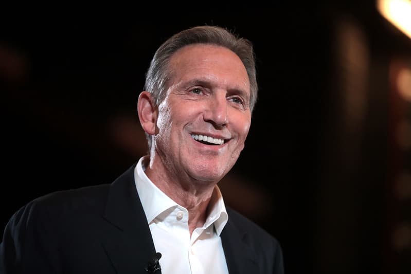 Howard Schultz wearing a dark blazer and a white shirt is standing in front of a blurred background. His expression conveys confidence and warmth, and he appears to be engaging with an audience or event.