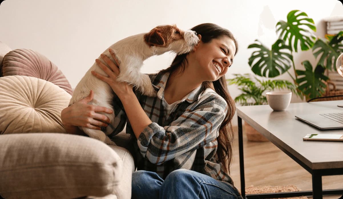 A woman is smiling and sitting on a sofa while holding a small dog that is playfully licking her face. She is wearing a plaid shirt and has long hair. The background features a home environment with a coffee table, a plant, and a laptop, creating a cozy atmosphere.