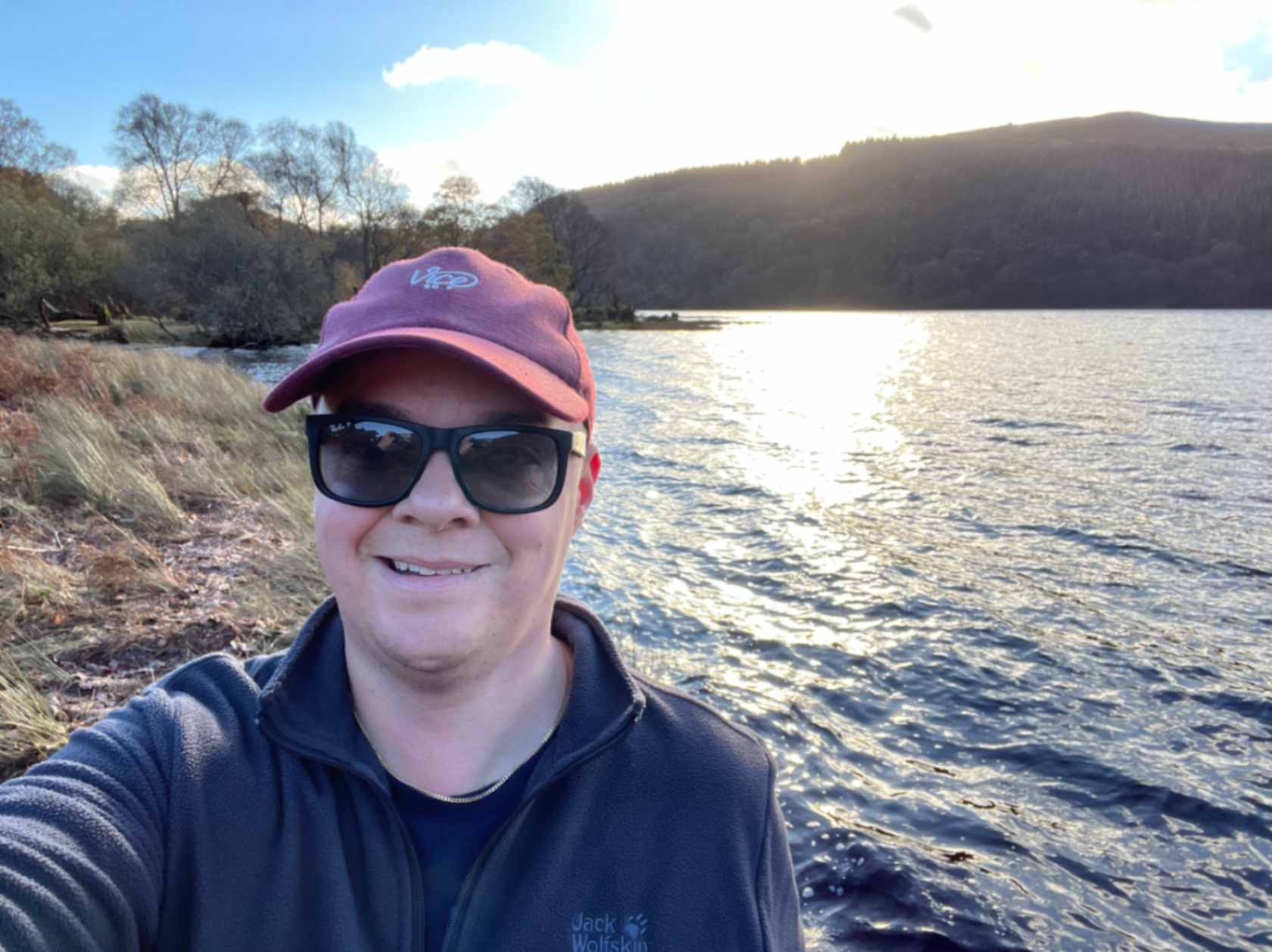 A person is smiling for a selfie by the edge of a body of water, wearing sunglasses and a maroon cap. In the background, there are trees and a hillside under a partly cloudy sky, with sunlight glistening on the water. The person is dressed casually in a dark sweater, and the setting suggests a tranquil outdoor environment.