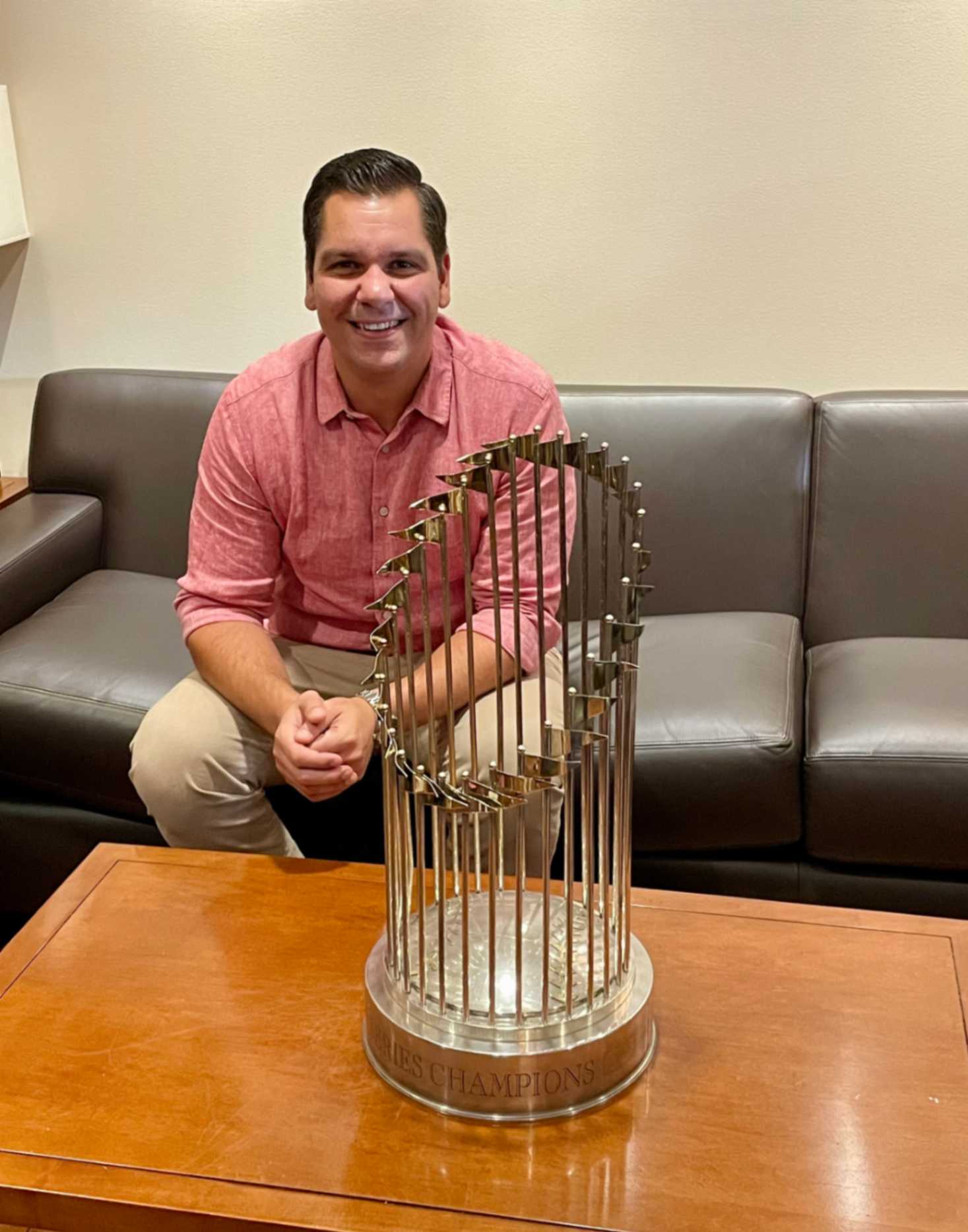 A man is smiling while sitting on a couch next to a championship trophy on a wooden table. The trophy features a shiny metallic design with vertical bars and has the text "CHAMPIONS" engraved on its base. The background includes a light-colored wall, enhancing the indoor setting.