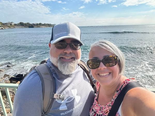 A smiling couple poses for a selfie near a body of water, with gentle waves in the background. The man is wearing a grey cap with "Workhuman" written on it and sunglasses, while the woman has short, grey hair and is wearing a colorful, floral-patterned shirt. The scene captures a sunny day with a clear sky.