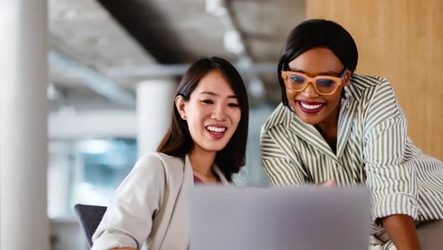 Two women are engaged in a conversation while looking at a laptop screen. One woman, wearing a light-colored blazer, is smiling and appears excited. The other woman, with glasses and wearing a striped top, is leaning over to point at something on the screen, also smiling. The background suggests an office setting with modern decor.