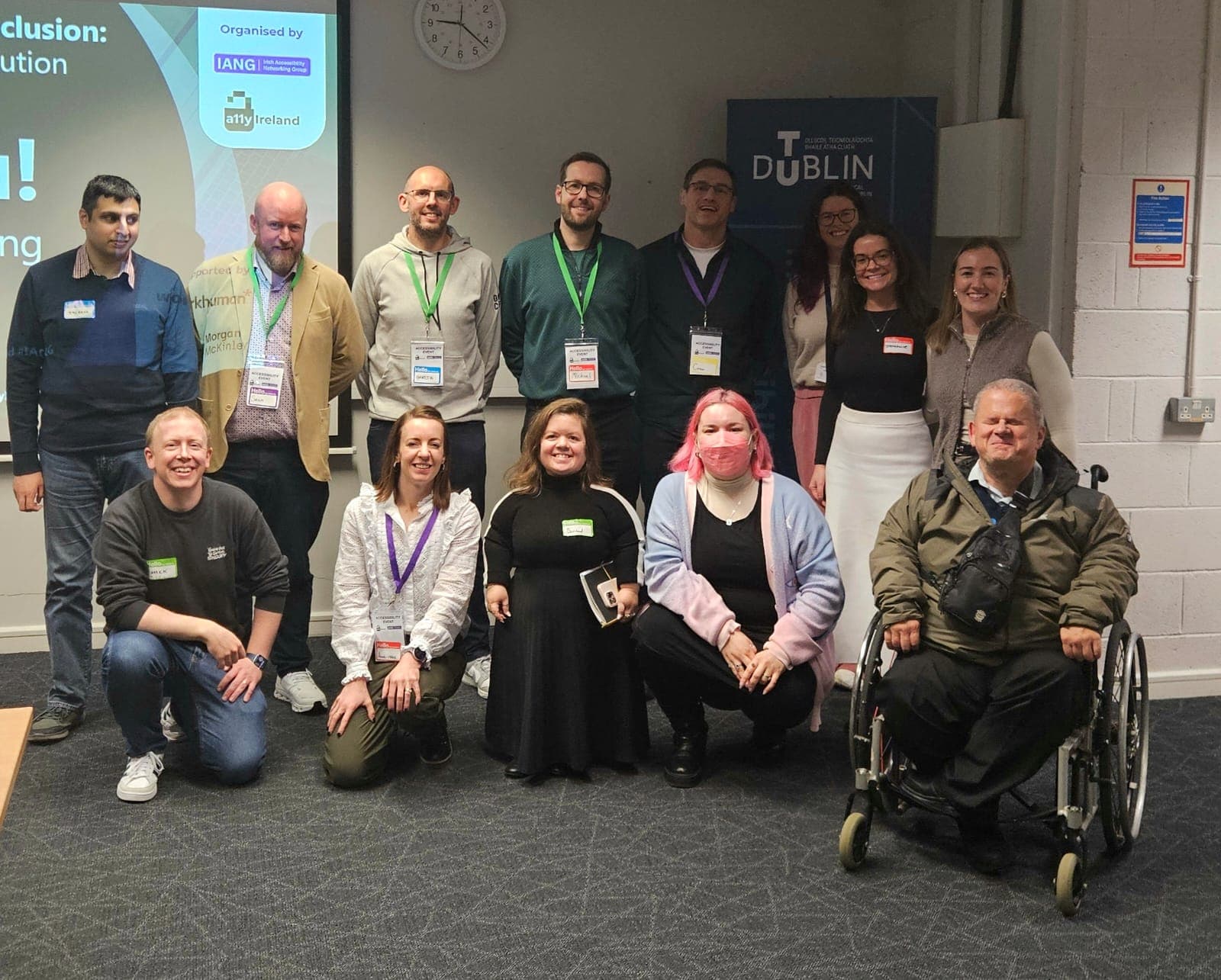 A group photo featuring attendees gathered for a conference on inclusion at a venue in Dublin. The group consists of multiple individuals, including those in wheelchairs and varying in height, showcasing diversity. They are standing and kneeling in front of a screen displaying the event’s title, with name tags visible on several participants. In the background, there’s a sign indicating the location. The overall atmosphere feels friendly and inclusive, with all individuals smiling.