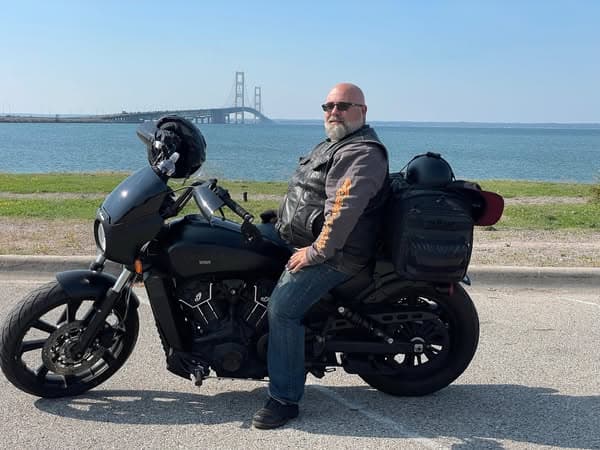 A man with a beard and sunglasses is sitting on a motorcycle parked by a waterfront. He is wearing a black leather jacket and blue jeans. In the background, a bridge spans across the water, under a clear blue sky. The scene captures a relaxed outdoor atmosphere.