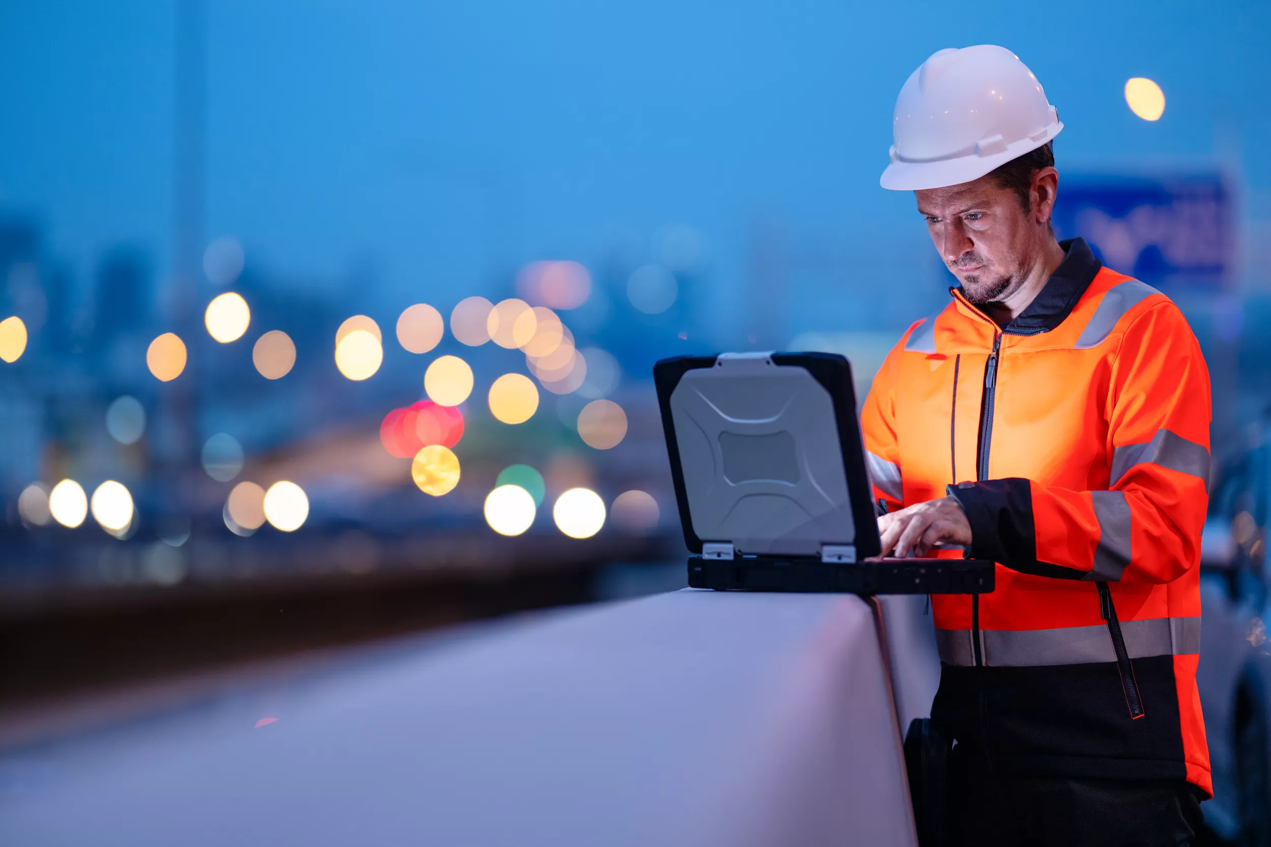 A worker in a safety vest and hard hat is using a laptop while standing by a railing in an urban setting during dusk. The background features blurred city lights, suggesting a busy street or highway illuminated with colorful bokeh effects. The worker appears focused on the laptop screen, with his attention directed towards the device.