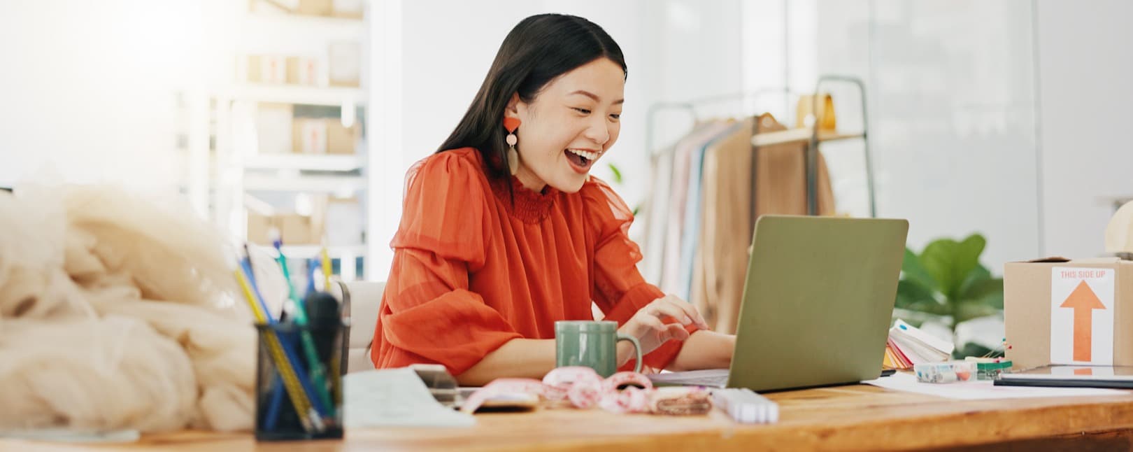 A woman in a red blouse is sitting at a wooden desk enthusiastically typing on a green laptop. She has long dark hair and is smiling widely. In front of her, there are a few colorful donuts, a green mug, and a container with pens and markers. The background shows shelves with boxes and clothing racks, creating a lively workspace atmosphere. The scene conveys a sense of joy and engagement in her work.