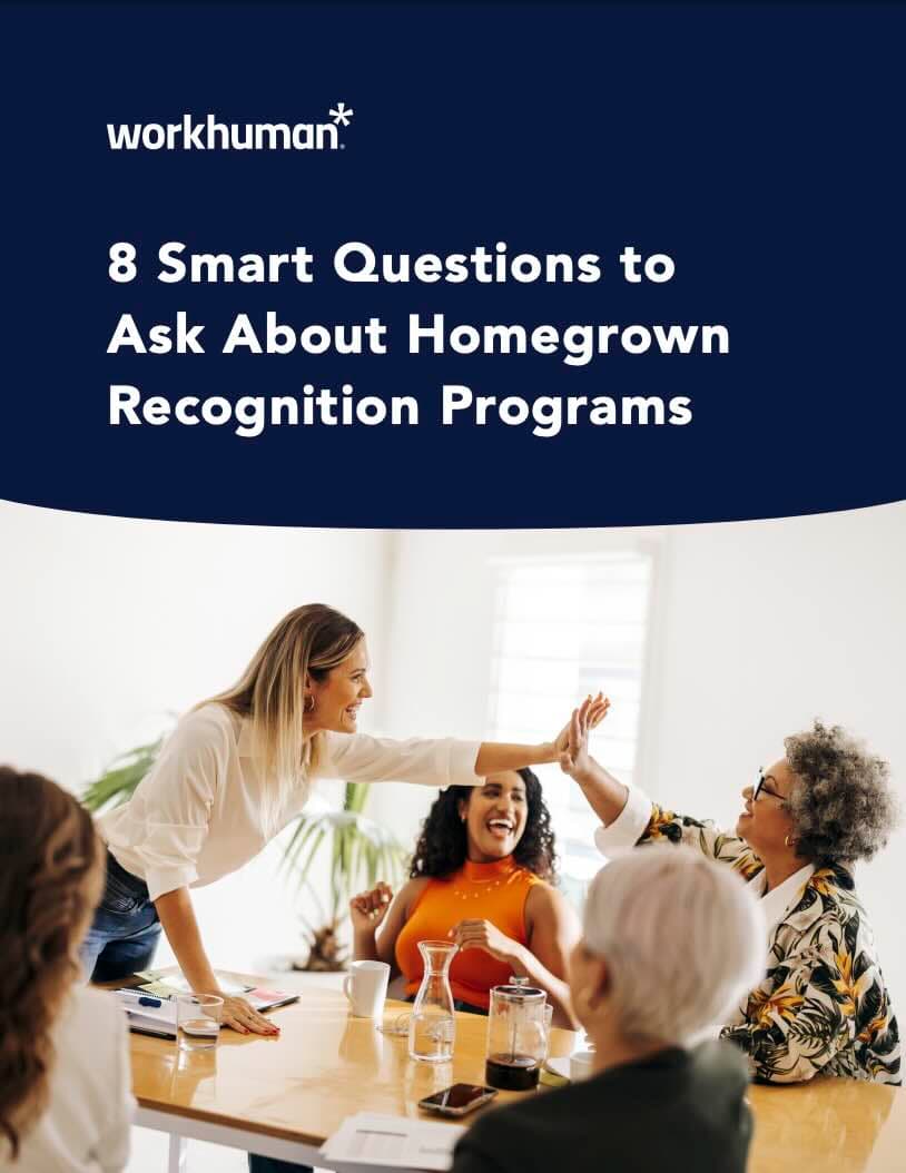 A group of four women are seated around a table in a bright, modern meeting room, engaged in a lively discussion. One woman, standing, is high-fiving another seated woman who appears to be laughing. The third woman, also seated, has a large smile, while the fourth woman, partially visible at the edge of the image, looks on. A pitcher and cups are positioned on the table, and there's a document and smartphone visible. The top of the image features the title: "8 Smart Questions to Ask About Homegrown Recognition Programs" alongside the Workhuman logo.