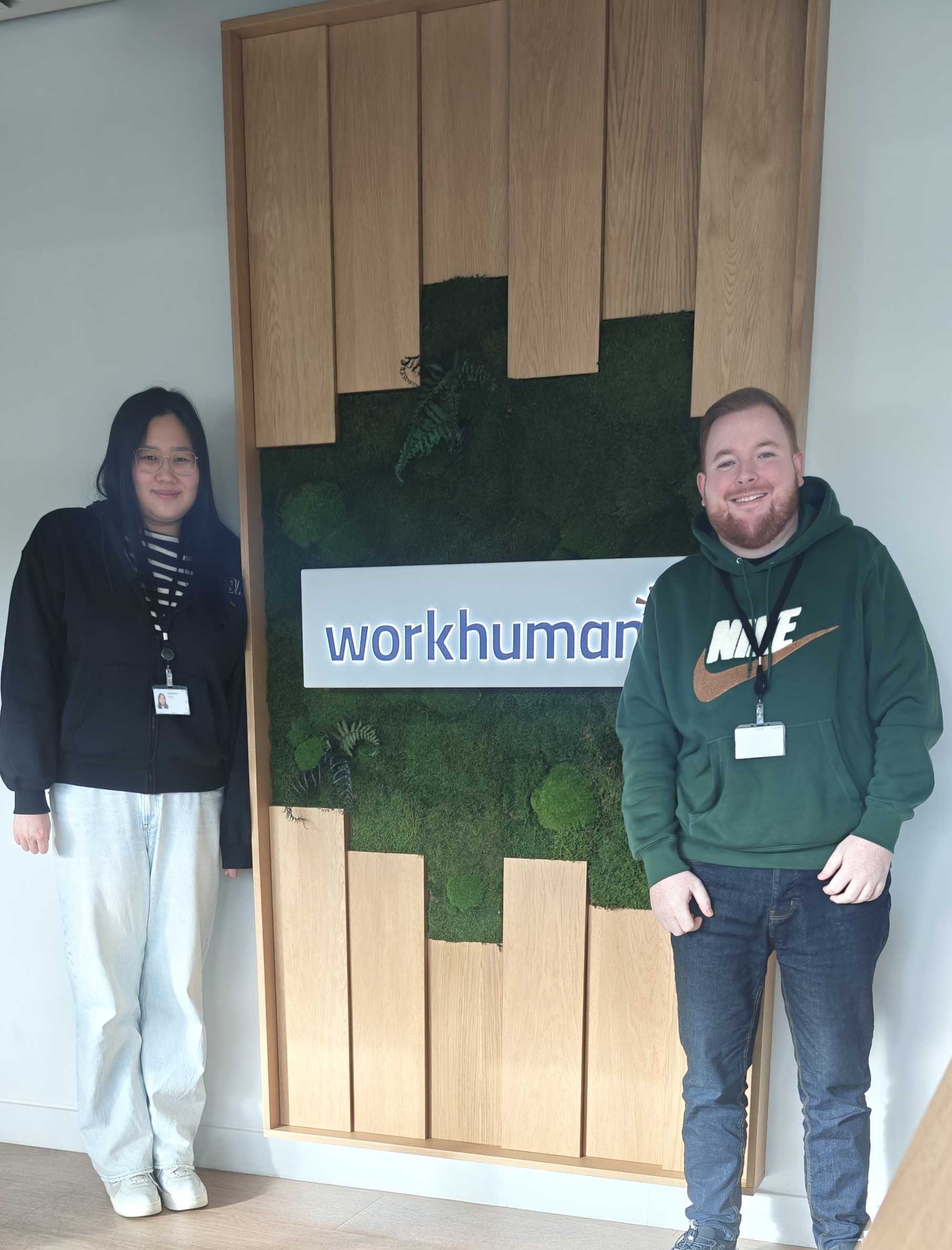 Two individuals are standing next to a decorative wall featuring the logo "workhuman." The backdrop consists of wooden panels with a green moss-like texture, enhancing the natural aesthetic of the space. One person, on the left, is wearing eyeglasses and a black jacket, while the other, on the right, is dressed in a green hoodie. Both are smiling, suggesting a friendly and welcoming environment.