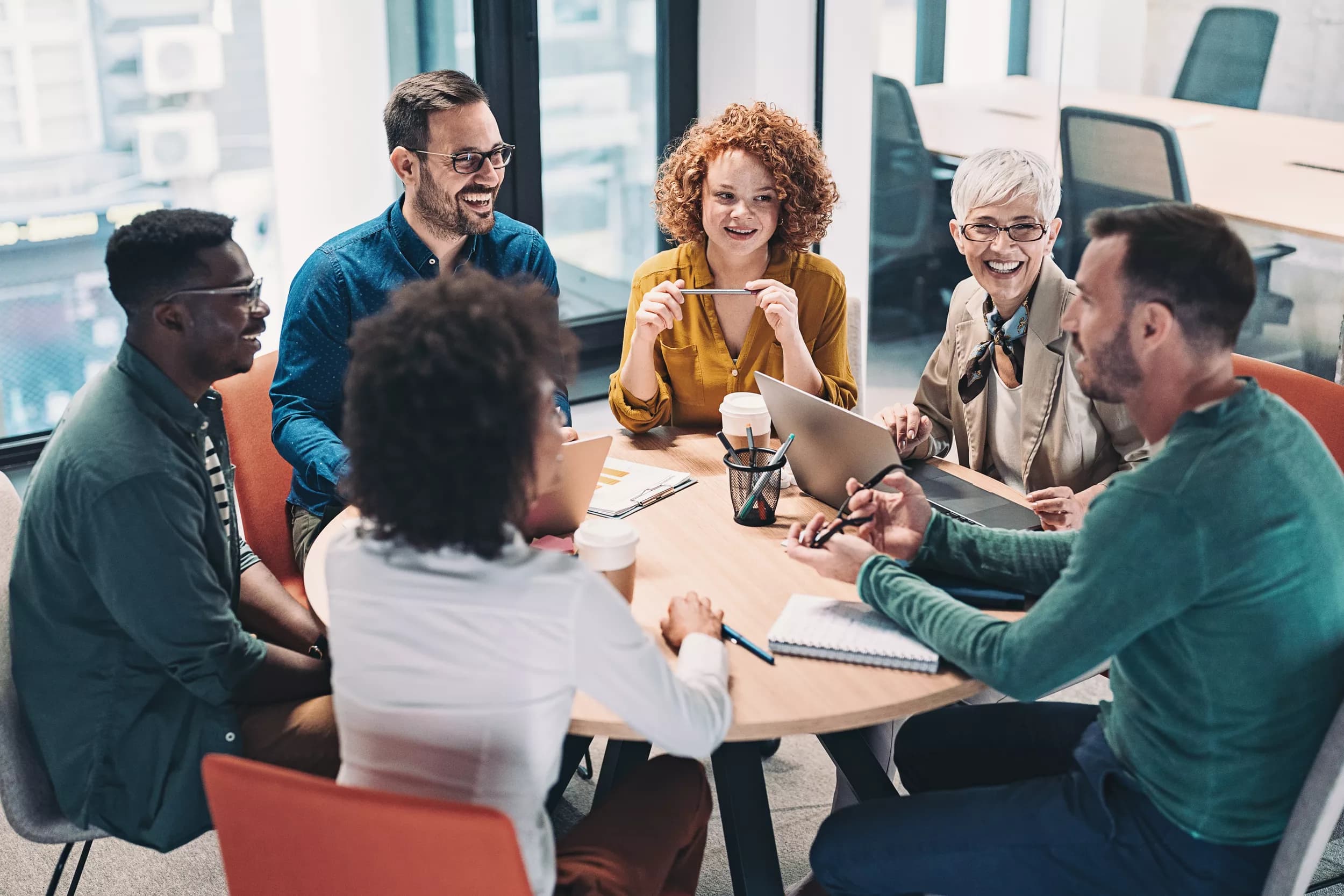 A diverse group of six colleagues sits around a circular table in a bright office setting for an agile team meeting. They appear engaged in conversation, with some smiling and laughing. Laptops and coffee cups are present on the table, indicating a collaborative work environment. The individuals are casually dressed, and the room has large windows that provide natural light, contributing to a warm and inviting atmosphere.