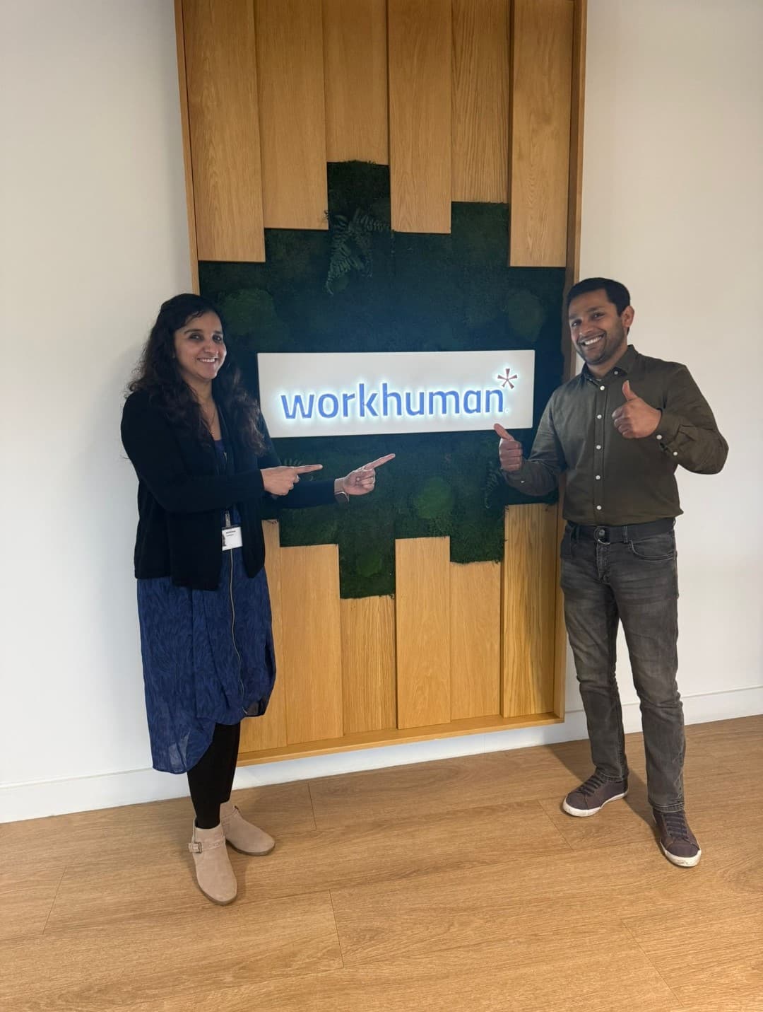 Two colleagues stand in an office environment in front of a sign that reads "workhuman." One person is pointing towards the sign while the other is giving a thumbs-up. The background features wooden panels and greenery, creating a welcoming atmosphere.