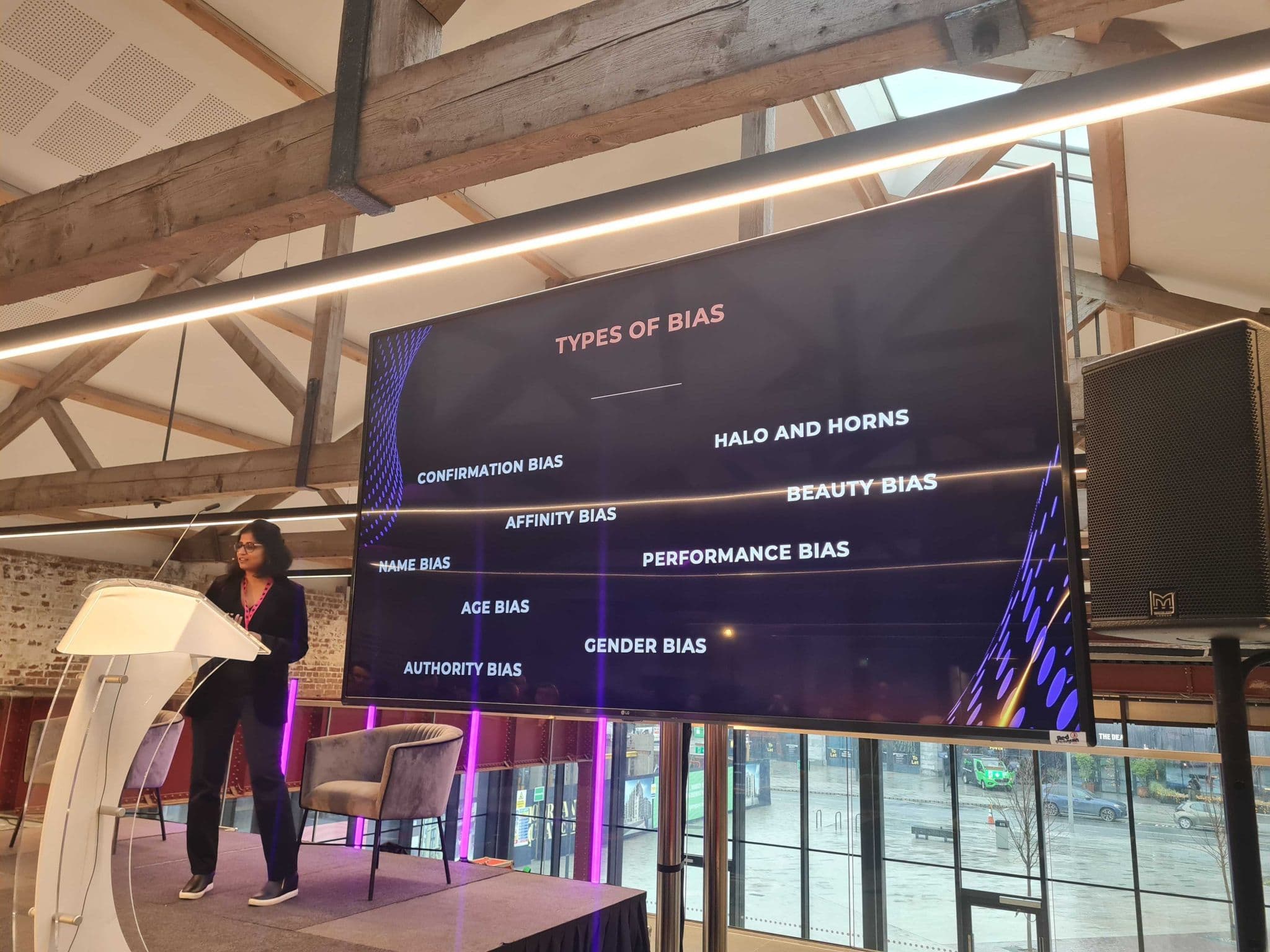 A woman standing on a stage presents a slide titled "TYPES OF BIAS," which lists various biases including Confirmation Bias, Affinity Bias, Name Bias, Age Bias, Gender Bias, Authority Bias, Halo and Horns, Beauty Bias, and Performance Bias. The setting features wooden beams overhead and modern furnishings, with a large screen displaying the information alongside a speaker podium.
