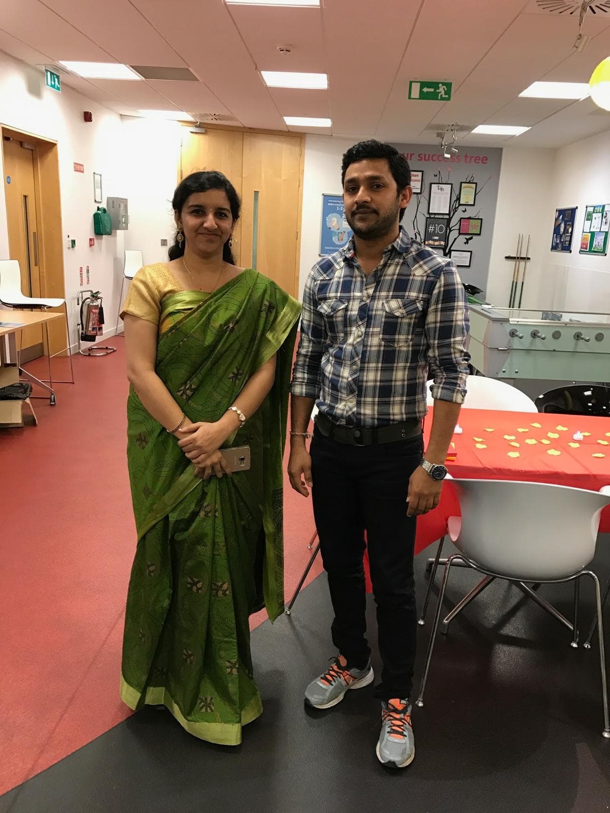 Two individuals are standing side by side in a well-lit office space. On the left, a woman is dressed in a green sari, holding a smartphone and smiling at the camera. On the right, a man in a plaid shirt and black pants stands with a neutral expression. In the background, there are desks, chairs, and a wall decorated with posters and a phrase that reads "our success tree." The floor has a reddish hue, and tables are adorned with small decorative items.