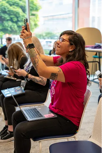 A woman with long dark hair and glasses is sitting on a chair, smiling as she takes a photo with her smartphone. She is wearing a bright pink shirt and has tattoos visible on her arms. In the background, several people are also seated, each looking at their devices. A laptop is open on her lap, and large windows allow sunlight to pour into the space, revealing greenery outside.
