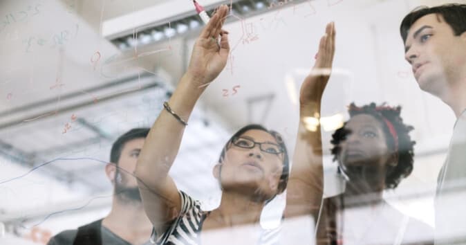 A diverse group of four people in a collaborative environment are engaged in a discussion, with one woman at the forefront using a marker to write on a transparent surface or whiteboard. The others are observing her, seemingly contributing to the conversation. The background includes various mathematical notations or diagrams that are faintly visible on the glass surface, indicating a brainstorming or planning session.