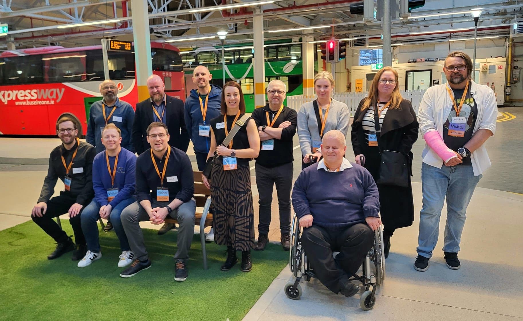 A group photo featuring fifteen individuals, including a man in a wheelchair, posing in a transportation setting. Some people are seated on a bench, while others stand behind them. Everyone wears visitor badges, and a red bus is visible in the background. The atmosphere appears friendly and casual.