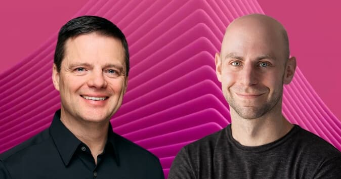 Two men are smiling and facing the camera, set against a vibrant pink background featuring undulating shapes. The man on the left has short dark hair and is wearing a black shirt, while the man on the right has a bald head and is dressed in a dark gray shirt. Both appear friendly and approachable.