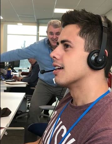 A young man is wearing a headset and focused on a task at a desk in a busy office environment, while an older man stands in the background, grinning and playfully reaching out towards him. The setting appears to be a call center or open office space, with several desks and coworkers visible in the background.