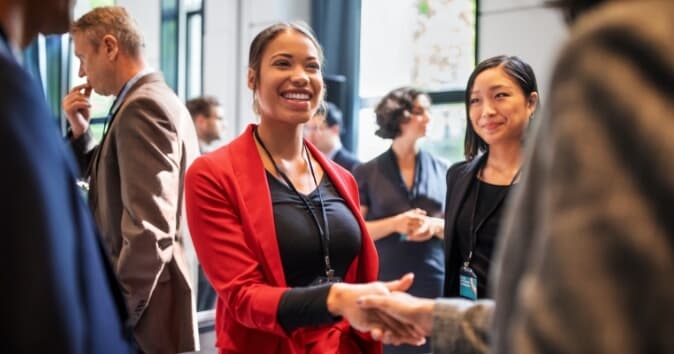 A smiling woman in a red blazer shakes hands with another person, engaging in conversation at a networking event. In the background, other attendees are seen interacting, with a focus on the social atmosphere of the gathering.