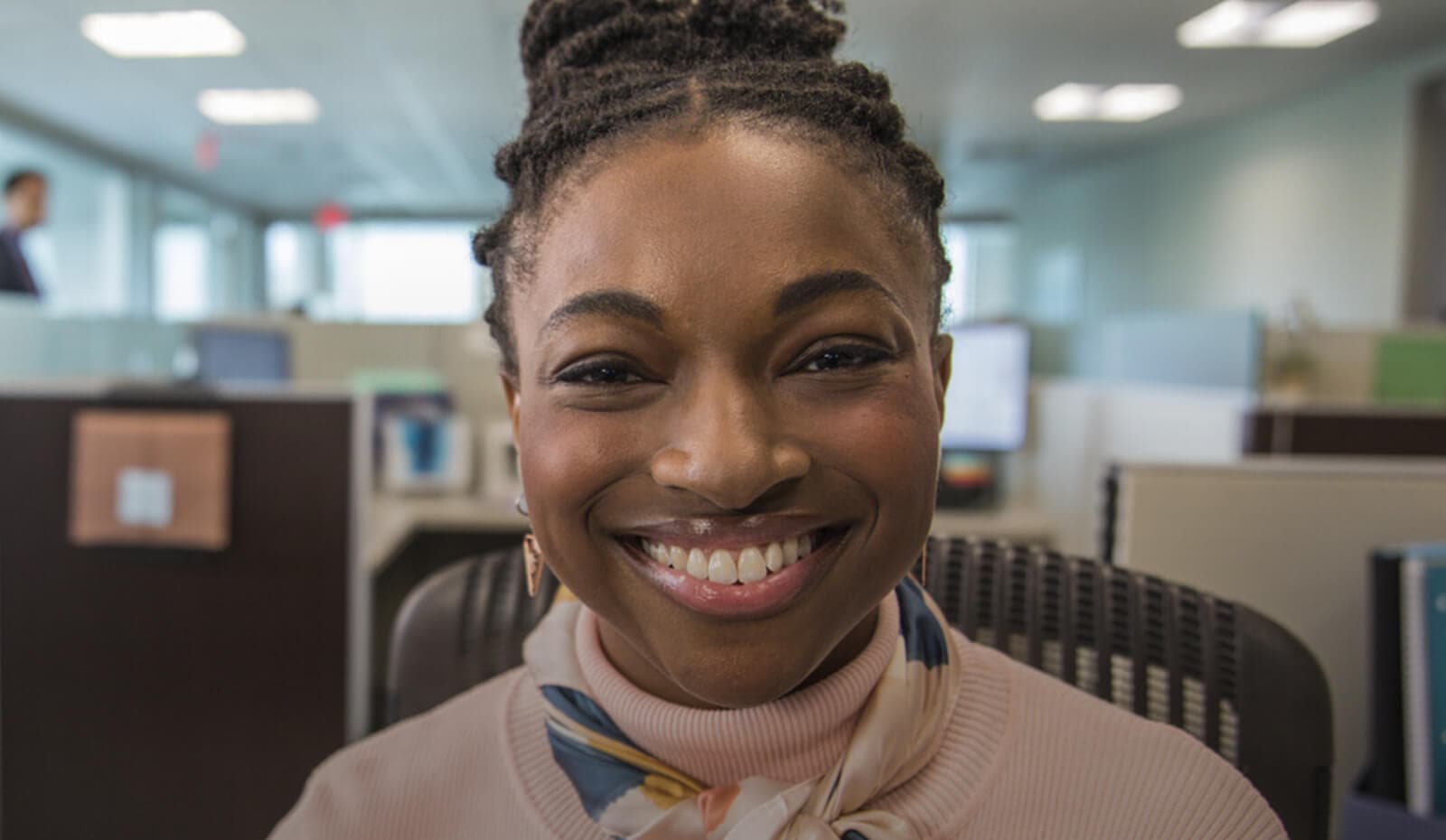 A young woman smiles at the camera while seated in an office environment, wearing a light pink sweater and a patterned scarf. The background shows cubicles and computer screens, suggesting a busy workplace atmosphere.