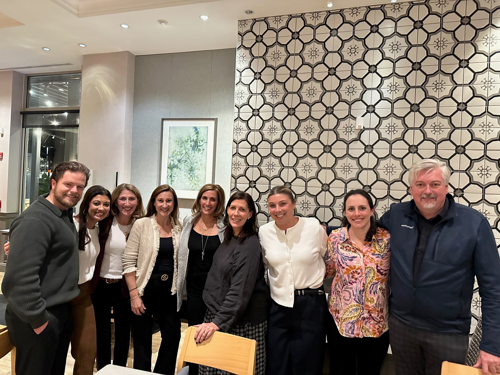 A group of nine people stands closely together in a warmly lit room with decorative patterned walls. They are smiling, conveying a sense of camaraderie.