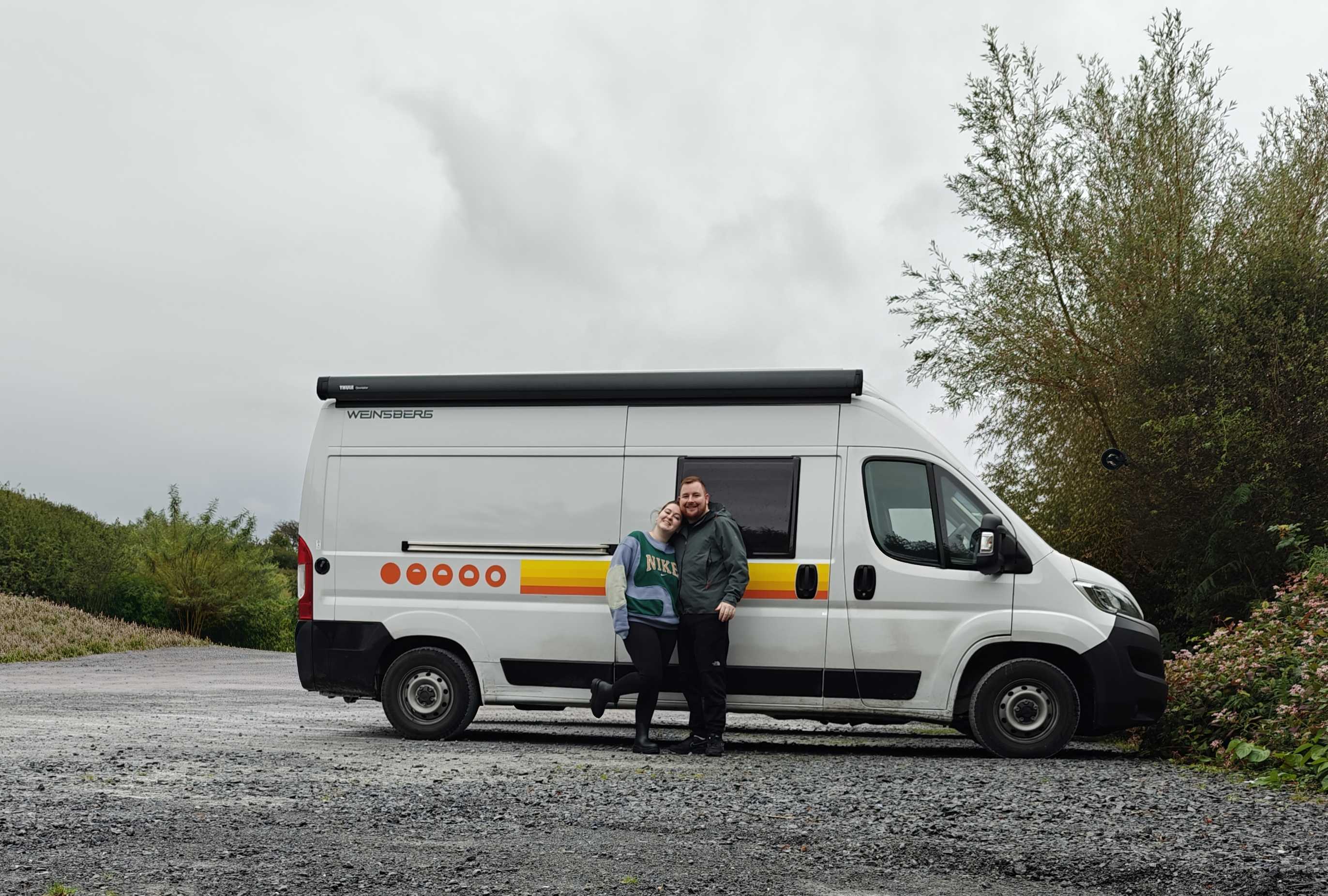 A couple stands smiling together next to a white camper van with a colorful stripe design. The van is parked on a gravel surface surrounded by greenery, and the sky appears cloudy. The woman is wearing a Nike sweatshirt, while the man is dressed in a green jacket.