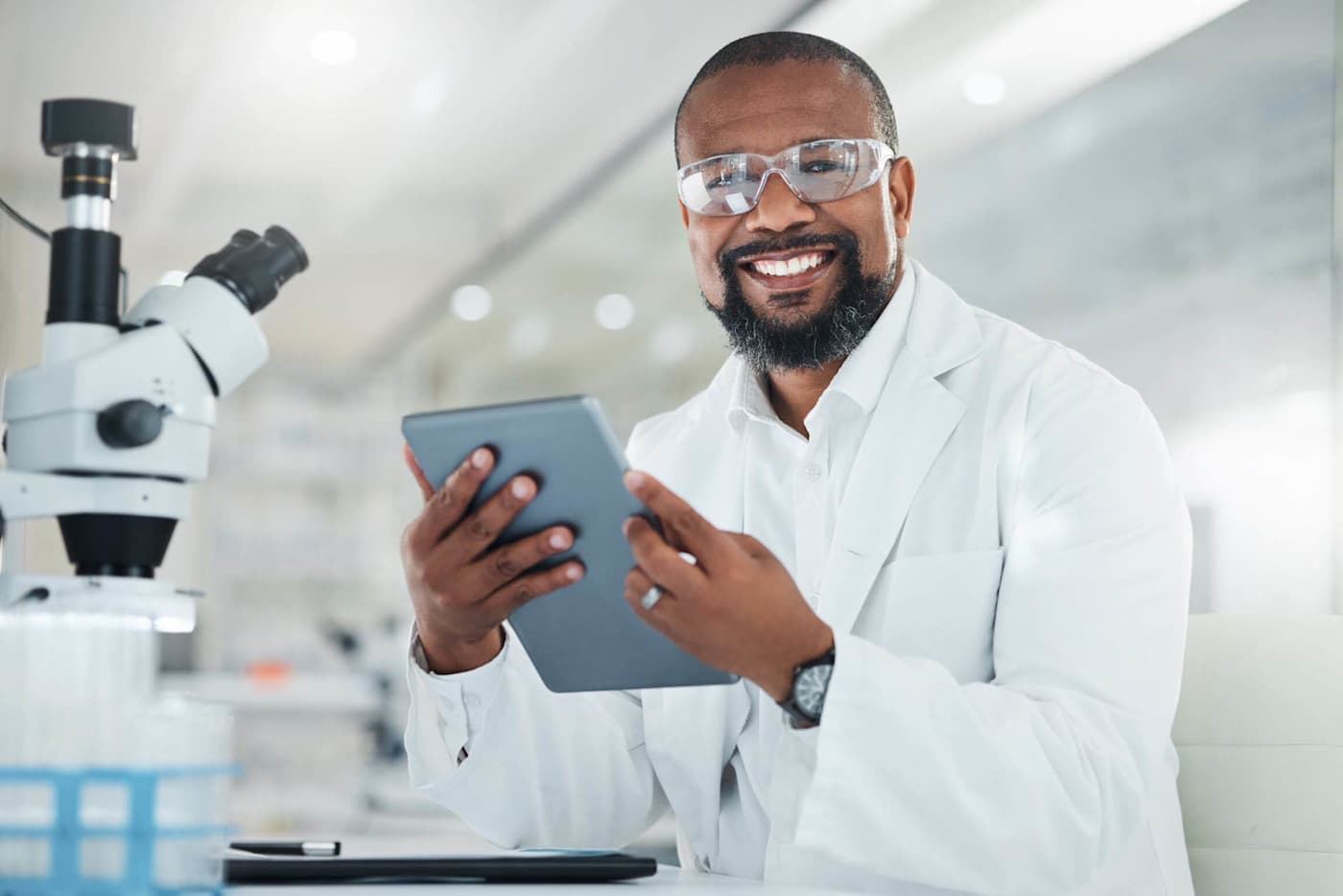 A person wearing protective eyewear and a lab coat is seated at a desk in a laboratory, holding a tablet and smiling. A microscope is visible on the left side of the image, with laboratory equipment in the background.