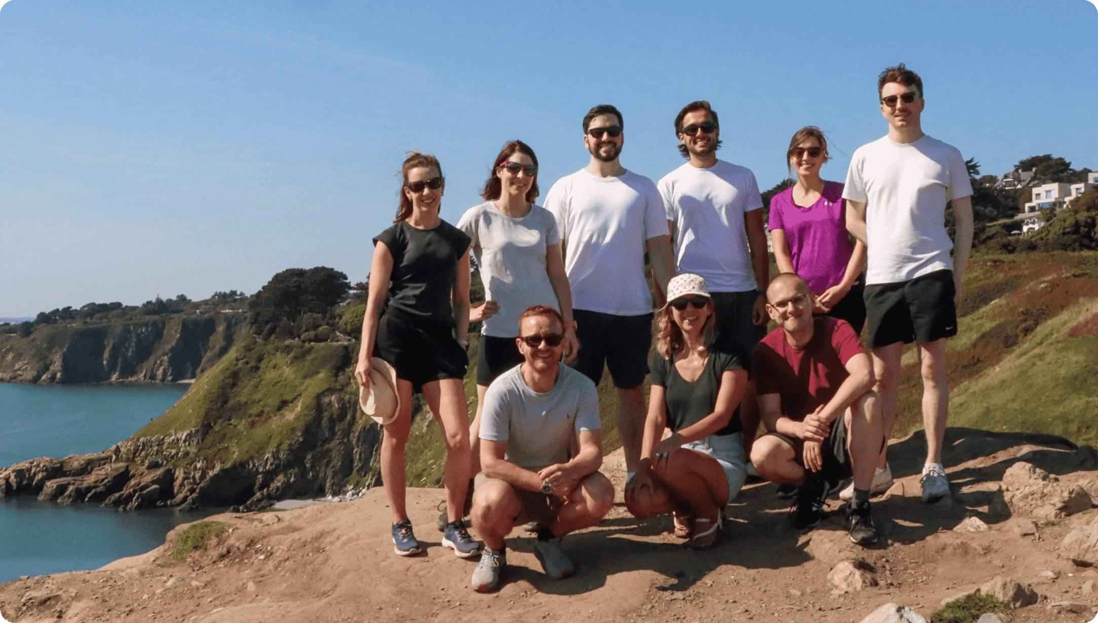9 members of the Terra team smile during a team day out on Howth hill. The team stand on a earthy mound with green cliffs jutting into the sea behind, and bright blue skies above