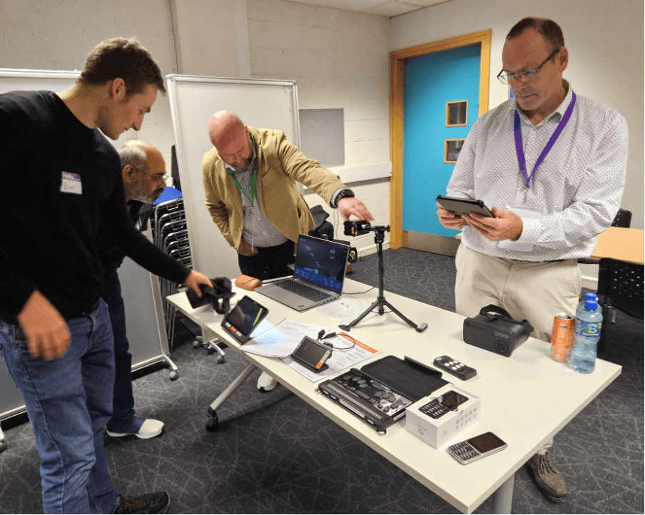 A group of four individuals is engaged in a hands-on discussion at a table laden with various electronic devices and tools. One person, wearing a black sweater with a name tag, is using a device while another, in a beige jacket, is pointing toward a camera mounted on a tripod. A third individual observes over the table, and a fourth is taking notes or using a tablet. Behind them, there's a doorway with blue panels and metal folding chairs stacked against the wall.