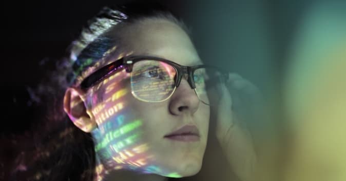A close-up of a woman wearing glasses, with a thoughtful expression as she appears to be looking at a projection or reflection of colorful text and patterns on her face. The lighting creates a vibrant effect, emphasizing the words and colors around her.
