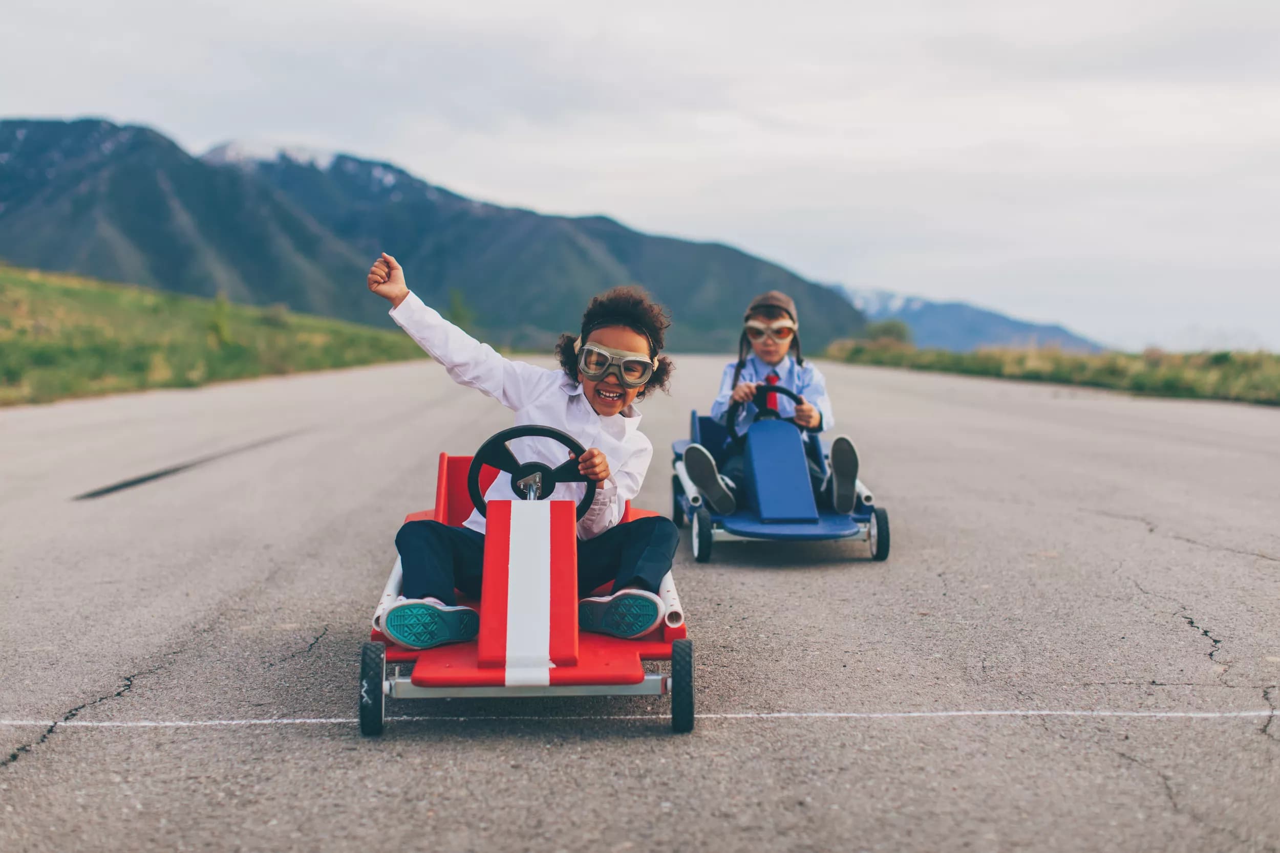 Two children are racing in go-karts along a smooth, paved track with mountains in the background. One child, in a red and white go-kart, is smiling and raising a fist in excitement, while the other child, in a blue go-kart, is focused on steering.