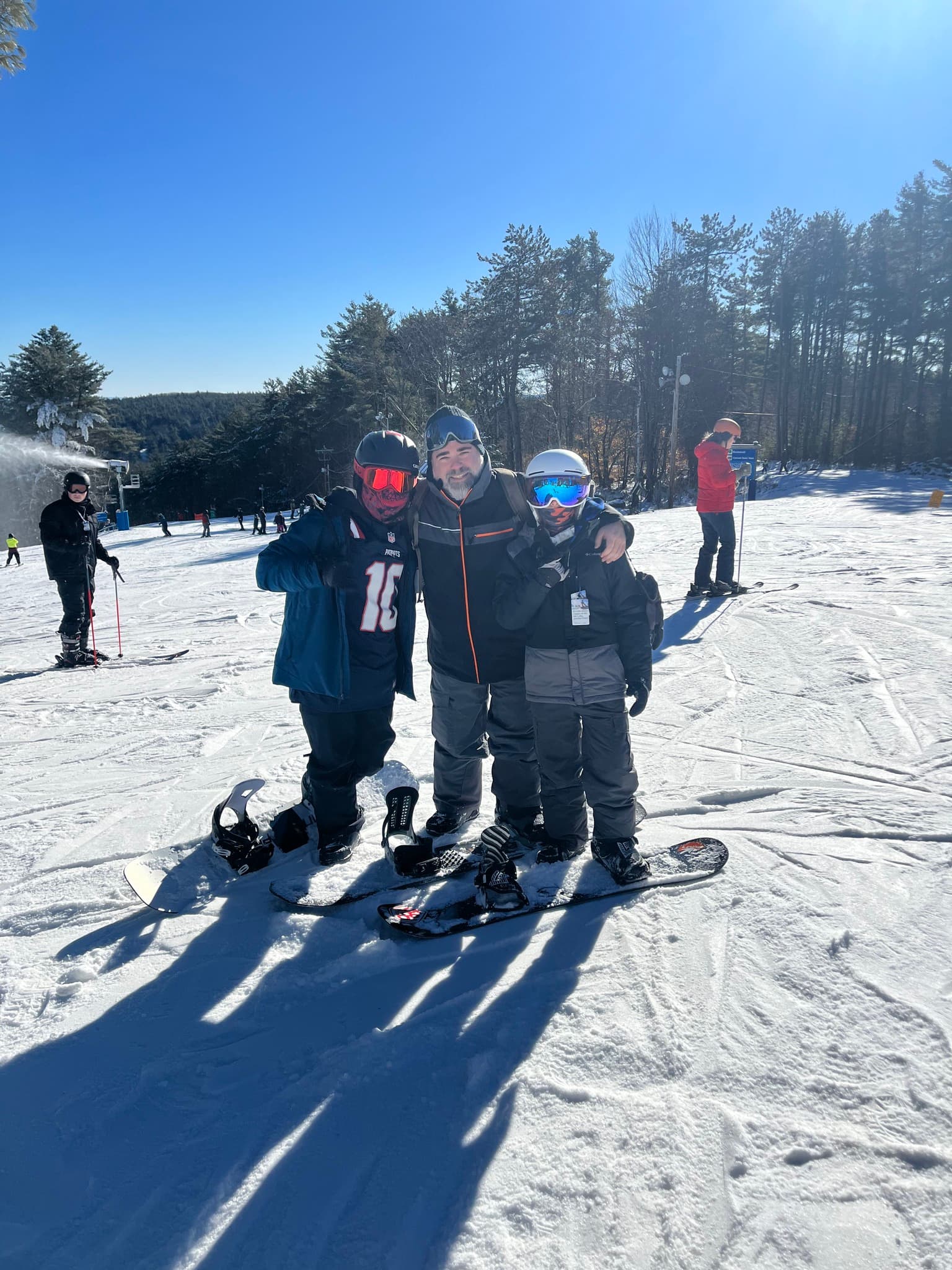 Three people stand on a snowy slope, smiling and posing together in ski gear. One person on the left wears a red jersey with the number "10" and has a face covering, while the person in the middle has a grey jacket and grey pants. The third person on the right sports a helmet and goggles. In the background, others ski in a winter landscape with trees and a blue sky. Snowboard equipment lies on the ground in front of them.