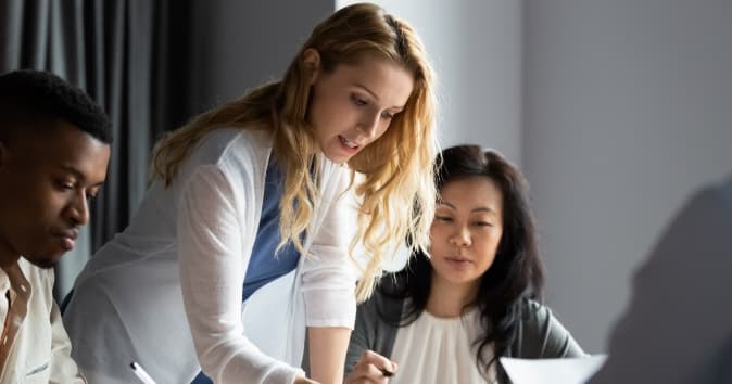 A collaborative work scene featuring three individuals engaged in a discussion at a table. A woman with long blonde hair is leaning over to assist, while two other individuals, one with short hair wearing a light-colored shirt and another with dark hair, are looking on attentively. Papers and writing materials are visible on the table, creating a focused and interactive atmosphere.