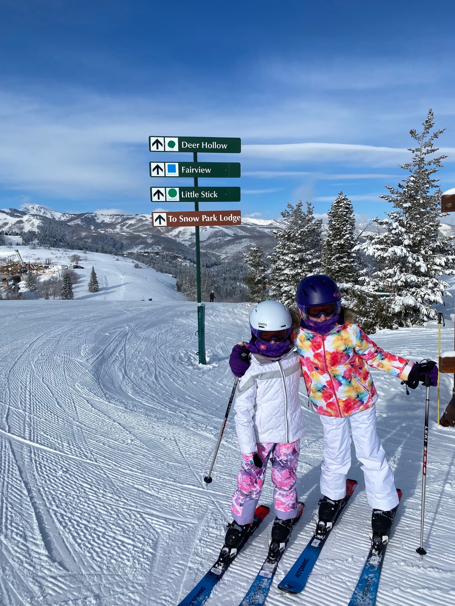 Two children in colorful ski gear stand on a snow-covered slope under a bright blue sky. A sign points to different ski trails with snowy mountains in the background.